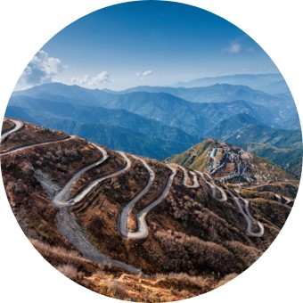 Das Bild zeigt Serpentinen in einer eher bräunlichen Berglandschaft. Dahinter sind blau-weiß Berge zu sehen und blauer Himmel mit wenigen Wolken.