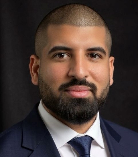 Porträt von Ali Bashirinia. Ein Mann mit 1mm kurzem schwarzen Haar und schwarzem Vollbart lächelt ohne Zähne in die Kamera. Er trägt eine dunkelblaue Anzugjacke, eine passende dunkelblaue Krawatte und ein weißes Hemd.