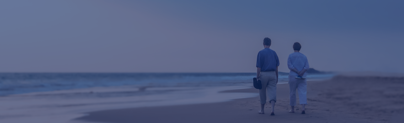 Zwei ältere Personen gehen barfuß entlang eines Strandes und tragen ihre Schuhe in der Hand, vor einer unscharfen Strandkulisse.