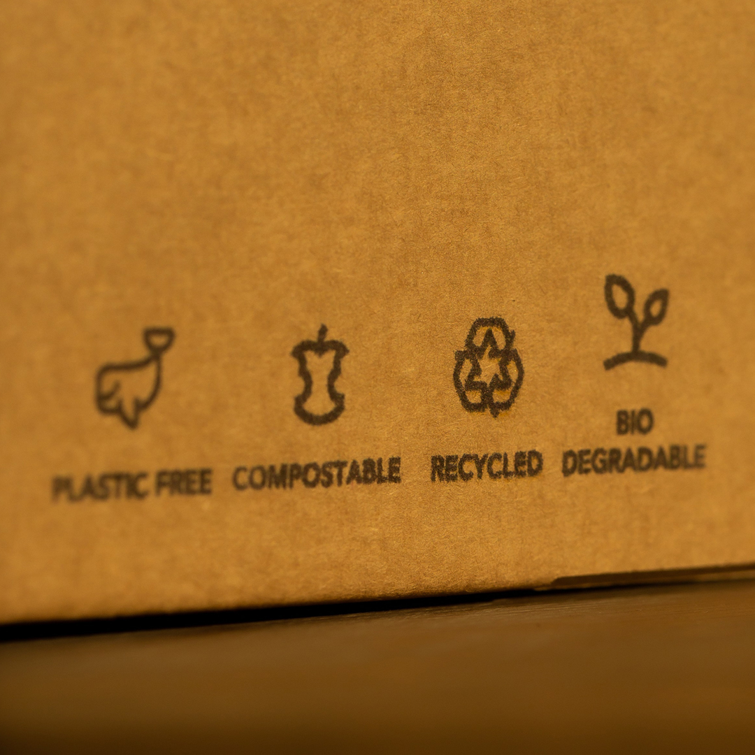 Auf einem Karton sind vier Symbole zu sehen: ein Wal mit der Unterschrift Plastikfrei, ein Apfelstrunk mit der Unterschrift Kompostierbar, drei Pfeile im Kreis mit der Unterschrift Recyclet und ein Setzling mit der Unterschrift Biologisch abbaubar - alle auf Englisch.